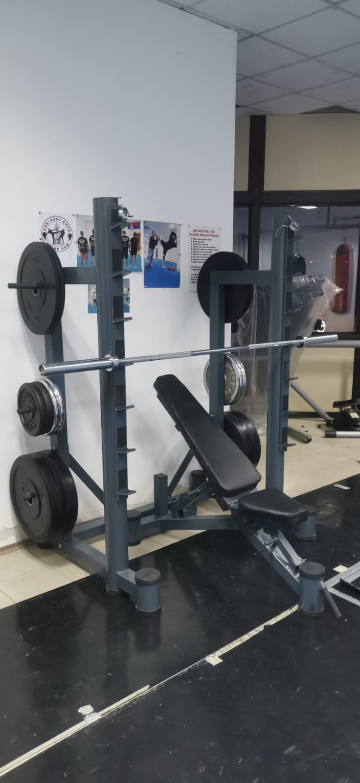 Stalak za čučanj sive boje sa trnovima za odlaganje tegova i kosom klupom u sklopu, u sali sportskog kluba